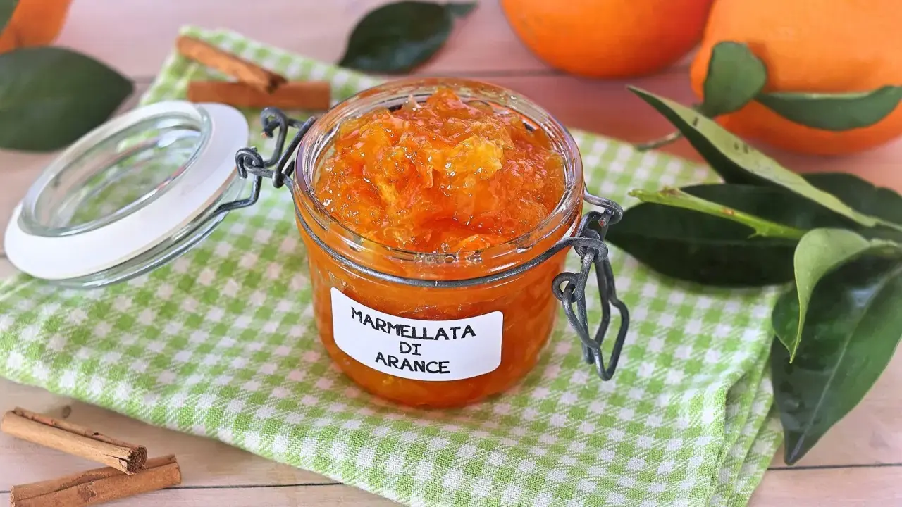 Barattolo di marmellata di arance fatto in casa, con arance fresche e bastoncini di cannella sullo sfondo.