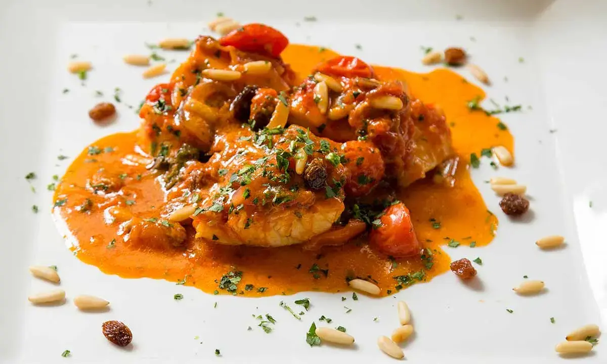 Un invitante piatto di baccalà alla romana, con pomodorini, pinoli e uvetta, immerso in una ricca salsa arancione e guarnito con prezzemolo tritato.