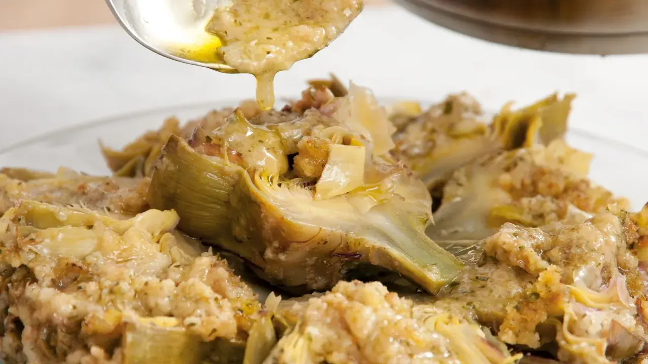 Un cucchiaio versa un sughetto dorato su un piatto di carciofi alla romana, un classico della cucina romana.