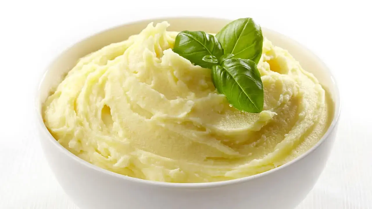 Una ciotola bianca piena di soffice crema di patate, guarnita con foglie di basilico fresco.