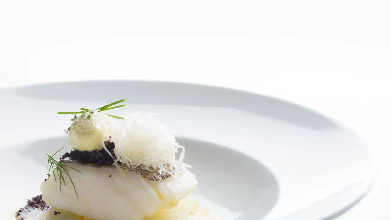 Piatto gourmet con filetto di pesce bianco, crema gialla, erbe aromatiche e fili croccanti. Un'interpretazione moderna che evoca la differenza tra baccalà e stoccafisso.