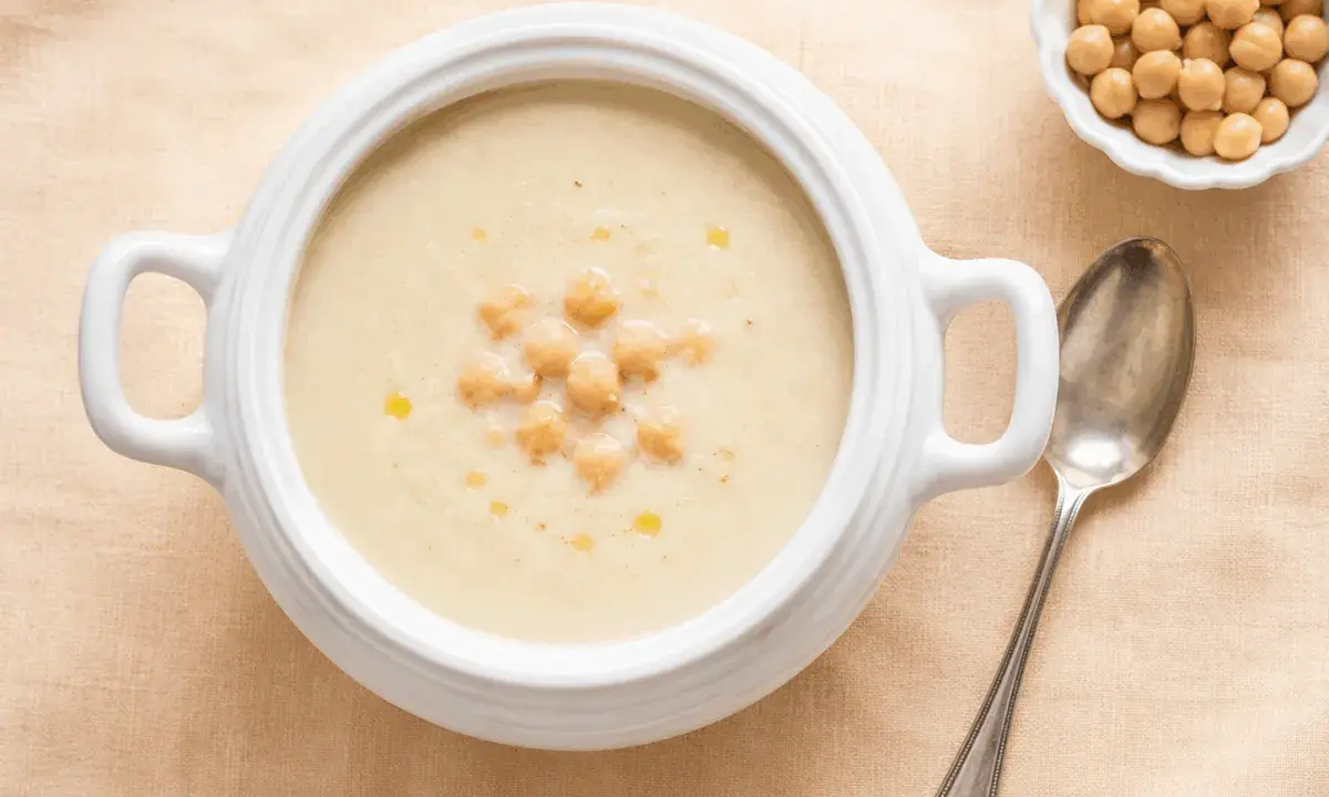 Zuppa cremosa di riso e ceci, guarnita con ceci interi e un filo d'olio. Accanto, una ciotolina di ceci e un cucchiaio d'argento.