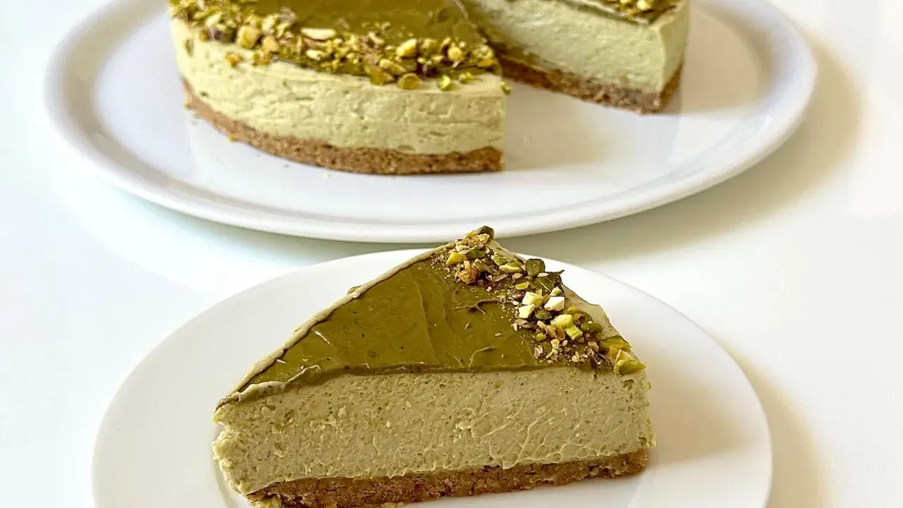 Una fetta di goloso cheesecake al pistacchio, con la torta intera sullo sfondo, decorata con granella di pistacchi.