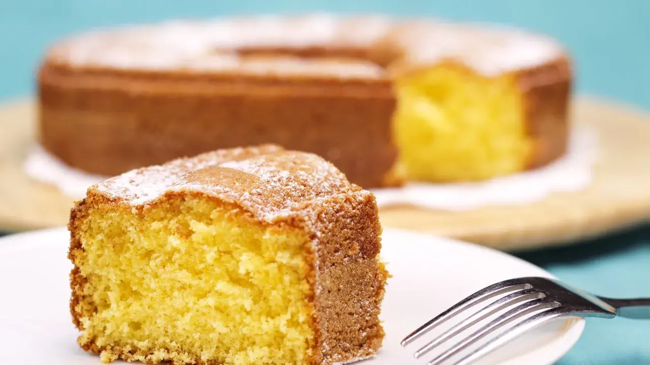 Una fetta di torta 7 vasetti soffice e dorata, cosparsa di zucchero a velo, su un piatto bianco con una forchetta.