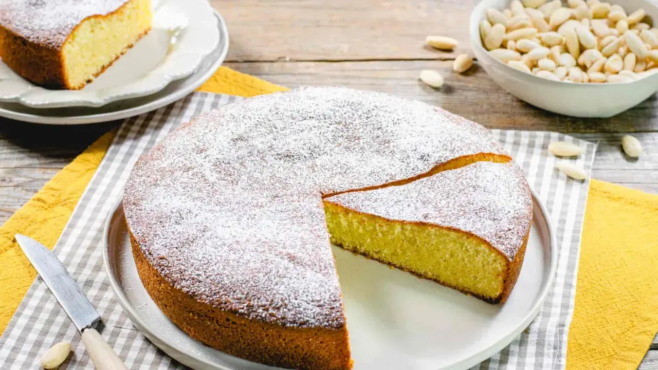 Una deliziosa torta con farina di mandorle, spolverata di zucchero a velo, pronta per essere gustata. Accanto, una ciotola di mandorle intere.