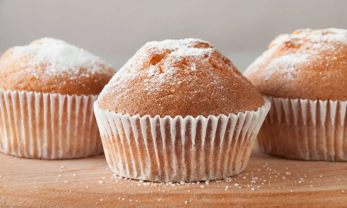 Tre deliziosi muffin senza glutine, spolverati di zucchero a velo, su un tagliere di legno.