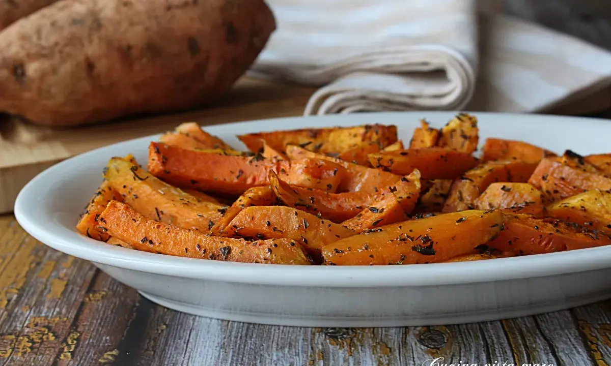 Patate dolci in friggitrice ad aria, tagliate a spicchi e condite con erbe aromatiche, servite in un piatto bianco.