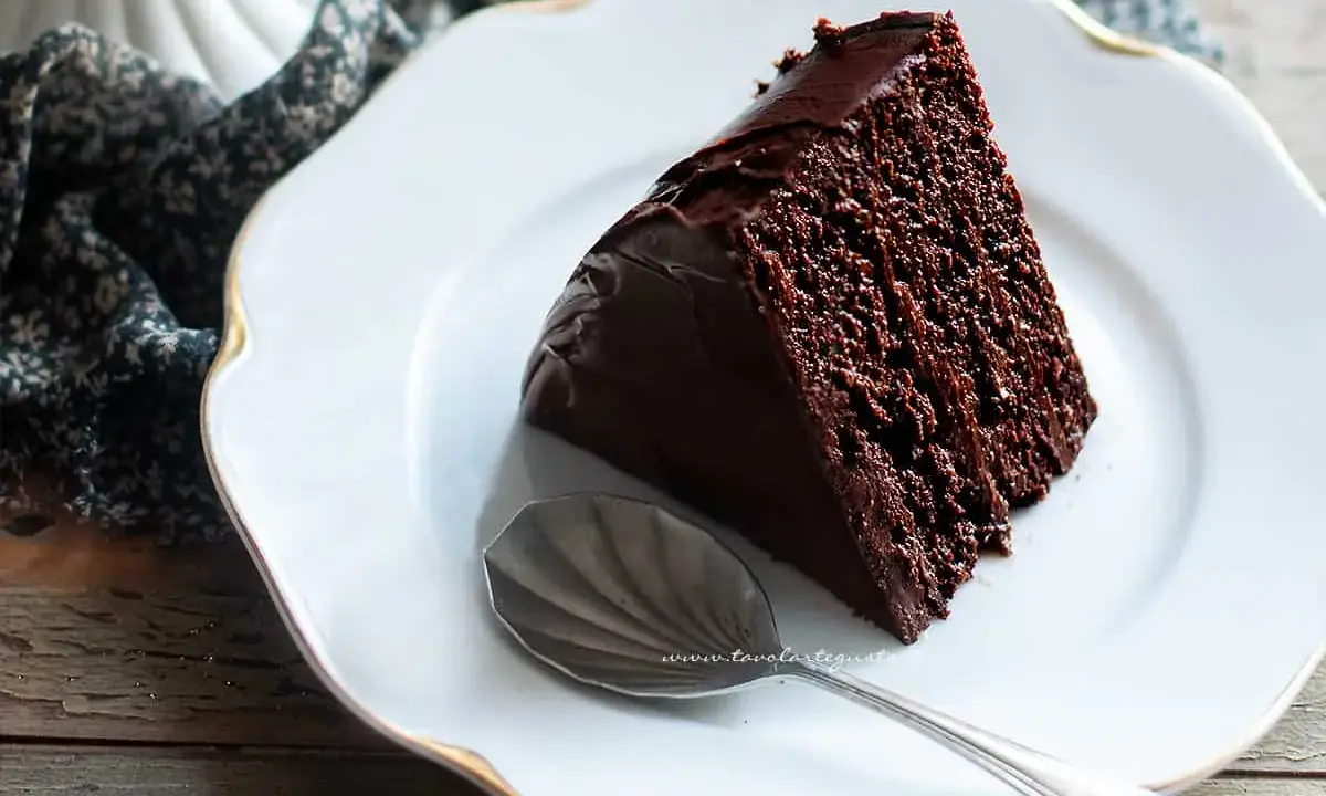 Una fetta di torta al cioccolato morbida e umida, ricoperta di glassa lucida, su un piatto bianco con un cucchiaino.