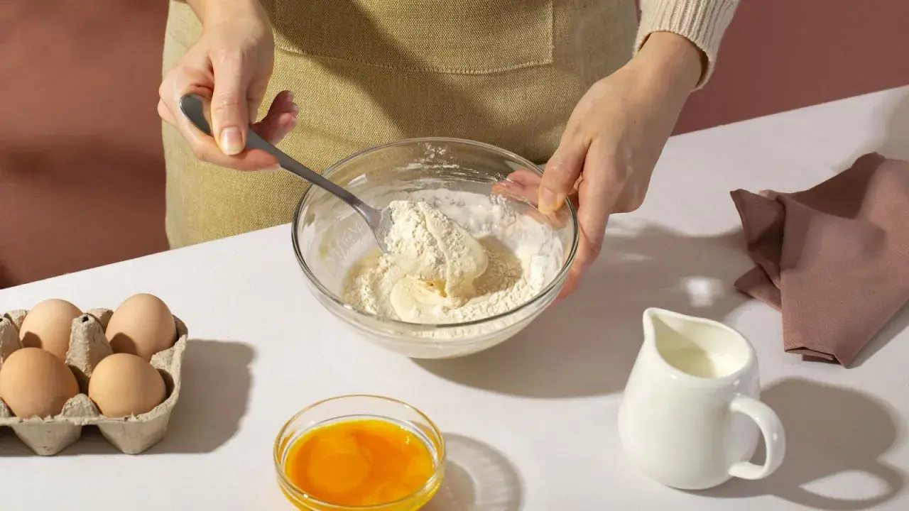 Mani che mescolano farina e burro in una ciotola, con uova, latte e bicarbonato al posto del lievito pronti per la preparazione.