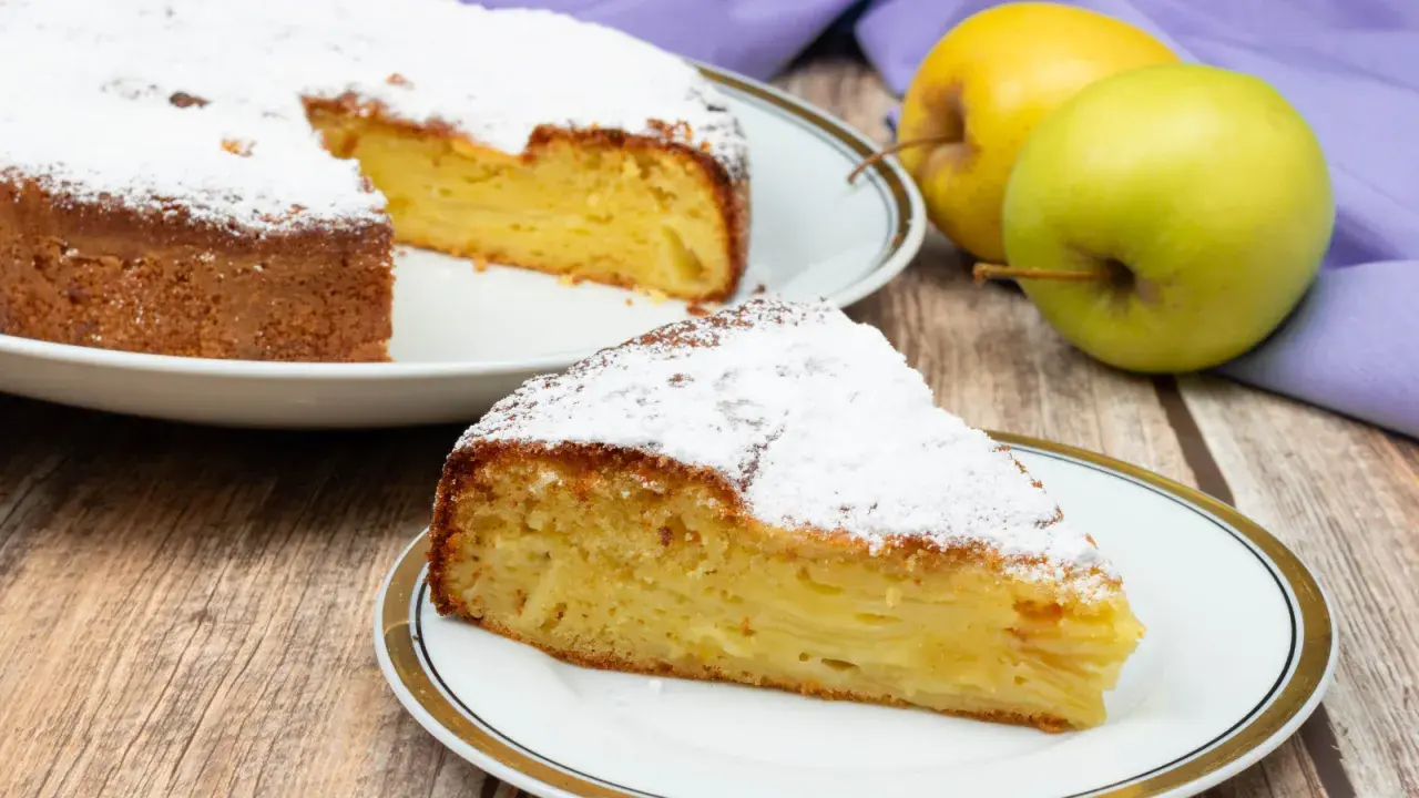 Una fetta di torta di mele invisibile, soffice e profumata, cosparsa di zucchero a velo. Accanto, mele fresche e un tovagliolo viola.