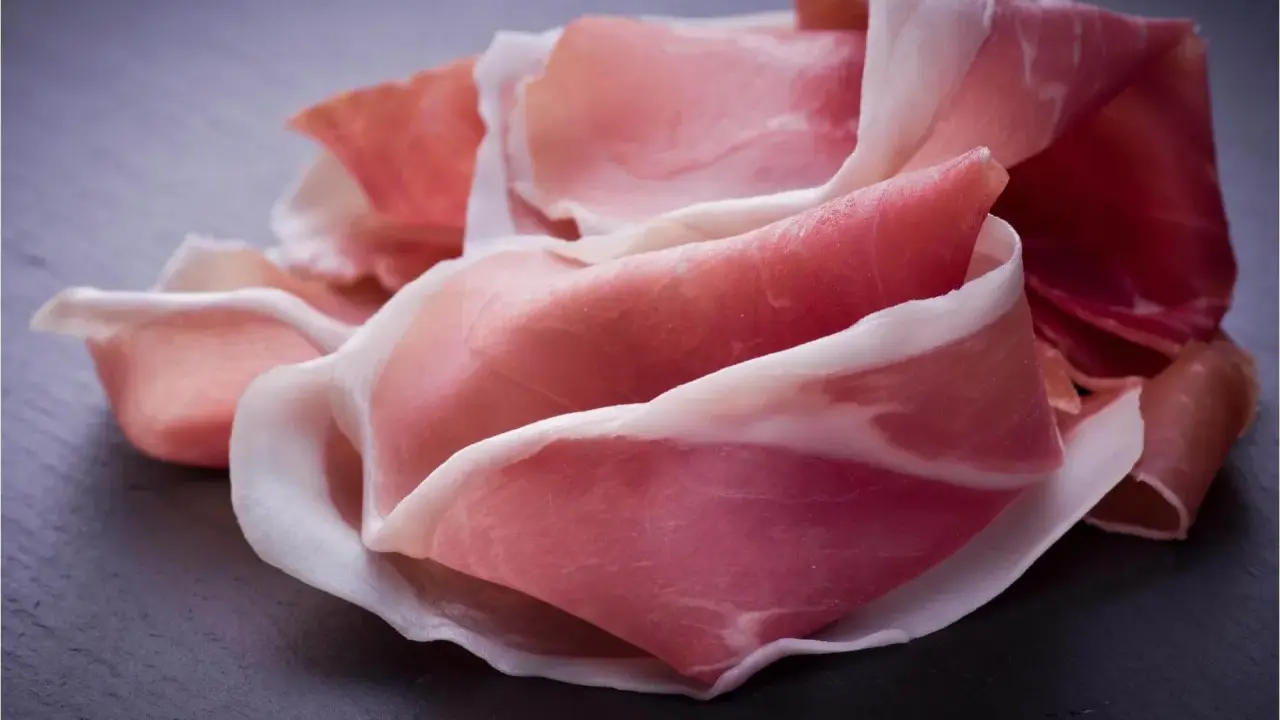 Fette di prosciutto crudo, un insaccato dal colore rosa intenso e venature bianche, disposte su una superficie scura.