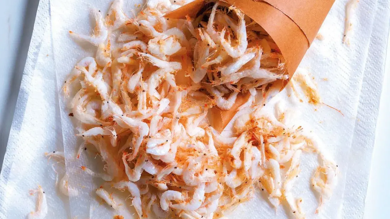 Piccolo cono di carta rovesciato su carta assorbente, con tanti gamberetti surgelati come cucinarli sparsi.