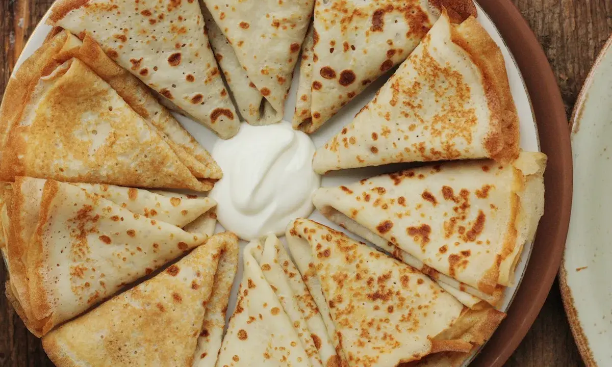 Piatto di crespelle ripiene, piegate a spicchi, con una generosa porzione di panna acida al centro.