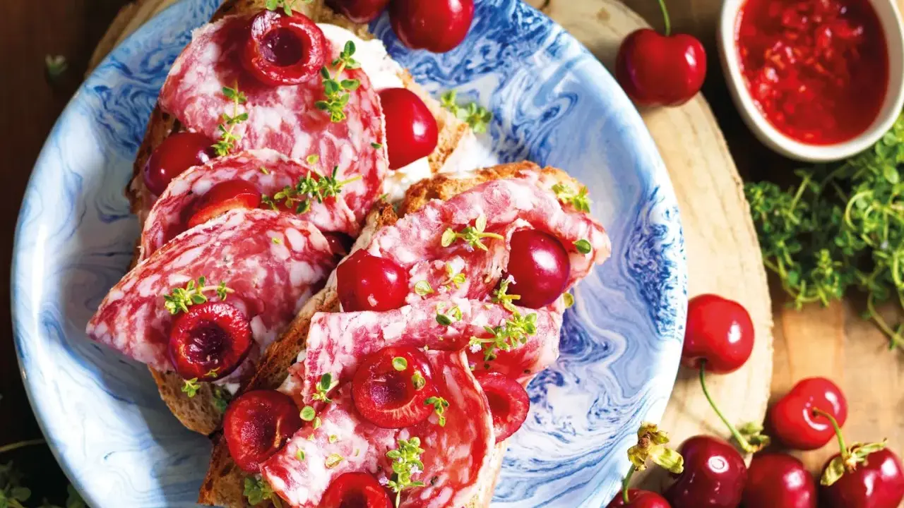 Bruschette con salame e varietà ciliegie giganti, guarnite con erbe aromatiche. Un connubio di sapori audace e invitante.
