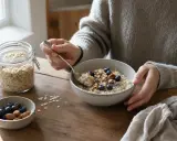 Persona che gusta una ciotola di fiocchi d'avena con mirtilli e noci. Un barattolo di fiocchi d'avena è visibile. Ecco come mangiare i fiocchi d'avena per una colazione sana.