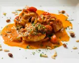 Un invitante piatto di baccalà alla romana, con pomodorini, pinoli e uvetta, immerso in una ricca salsa arancione e guarnito con prezzemolo tritato.