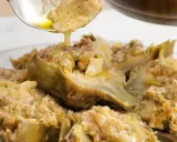 Un cucchiaio versa un sughetto dorato su un piatto di carciofi alla romana, un classico della cucina romana.