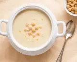 Zuppa cremosa di riso e ceci, guarnita con ceci interi e un filo d'olio. Accanto, una ciotolina di ceci e un cucchiaio d'argento.