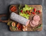 Assortimento di tipi di salame su tagliere in legno, con pomodorini, lattuga e fichi.
