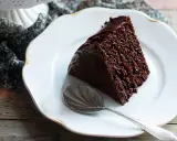 Una fetta di torta al cioccolato morbida e umida, ricoperta di glassa lucida, su un piatto bianco con un cucchiaino.