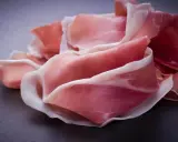 Fette di prosciutto crudo, un insaccato dal colore rosa intenso e venature bianche, disposte su una superficie scura.