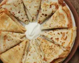 Piatto di crespelle ripiene, piegate a spicchi, con una generosa porzione di panna acida al centro.