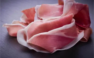 Fette di prosciutto crudo, un insaccato dal colore rosa intenso e venature bianche, disposte su una superficie scura.