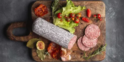 Assortimento di tipi di salame su tagliere in legno, con pomodorini, lattuga e fichi.