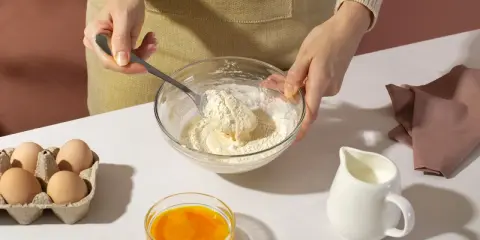 Mani che mescolano farina e burro in una ciotola, con uova, latte e bicarbonato al posto del lievito pronti per la preparazione.
