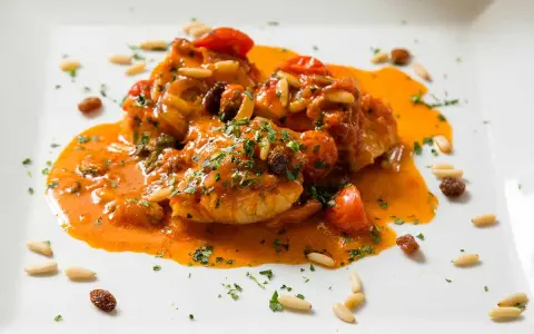 Un invitante piatto di baccalà alla romana, con pomodorini, pinoli e uvetta, immerso in una ricca salsa arancione e guarnito con prezzemolo tritato.