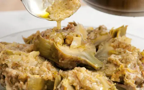 Un cucchiaio versa un sughetto dorato su un piatto di carciofi alla romana, un classico della cucina romana.