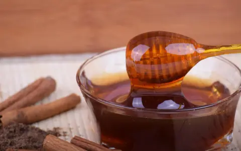 Miele di castagno, ricco di proprietà benefiche, gocciola da un cucchiaio in una ciotola. Cannella e polvere scura completano la scena.