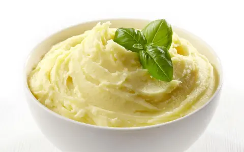 Una ciotola bianca piena di soffice crema di patate, guarnita con foglie di basilico fresco.