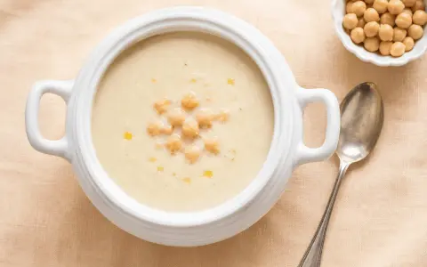 Zuppa cremosa di riso e ceci, guarnita con ceci interi e un filo d'olio. Accanto, una ciotolina di ceci e un cucchiaio d'argento.
