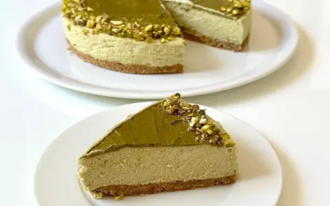Una fetta di goloso cheesecake al pistacchio, con la torta intera sullo sfondo, decorata con granella di pistacchi.
