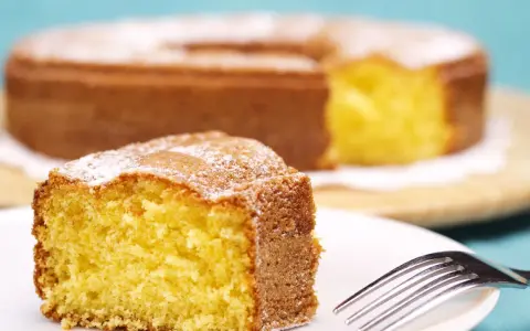 Una fetta di torta 7 vasetti soffice e dorata, cosparsa di zucchero a velo, su un piatto bianco con una forchetta.