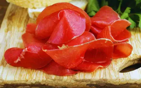 Fette sottili di bresaola, un insaccato di manzo stagionato, disposte su un tagliere di legno con pane e prezzemolo sullo sfondo.