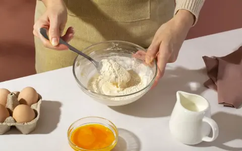 Mani che mescolano farina e burro in una ciotola, con uova, latte e bicarbonato al posto del lievito pronti per la preparazione.