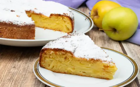 Una fetta di torta di mele invisibile, soffice e profumata, cosparsa di zucchero a velo. Accanto, mele fresche e un tovagliolo viola.