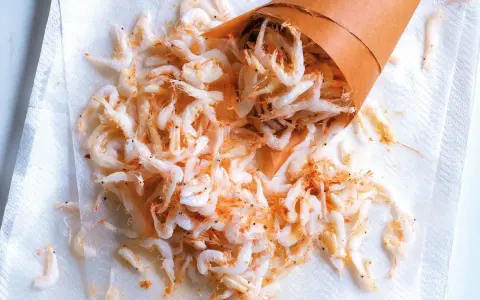 Piccolo cono di carta rovesciato su carta assorbente, con tanti gamberetti surgelati come cucinarli sparsi.