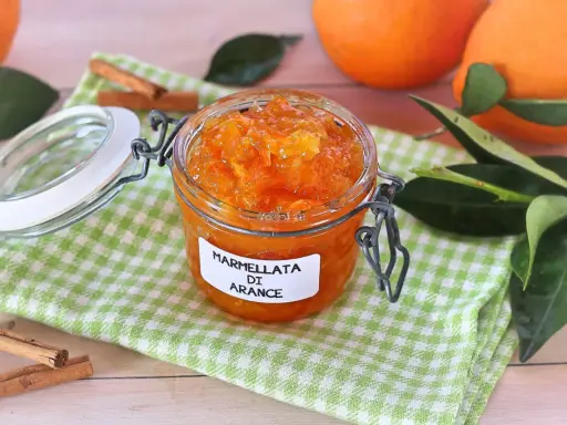 Barattolo di marmellata di arance fatto in casa, con arance fresche e bastoncini di cannella sullo sfondo.
