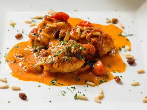 Un invitante piatto di baccalà alla romana, con pomodorini, pinoli e uvetta, immerso in una ricca salsa arancione e guarnito con prezzemolo tritato.
