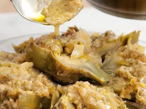 Un cucchiaio versa un sughetto dorato su un piatto di carciofi alla romana, un classico della cucina romana.