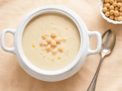 Zuppa cremosa di riso e ceci, guarnita con ceci interi e un filo d'olio. Accanto, una ciotolina di ceci e un cucchiaio d'argento.