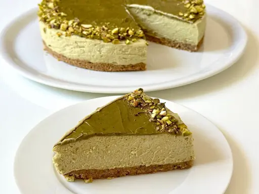 Una fetta di goloso cheesecake al pistacchio, con la torta intera sullo sfondo, decorata con granella di pistacchi.