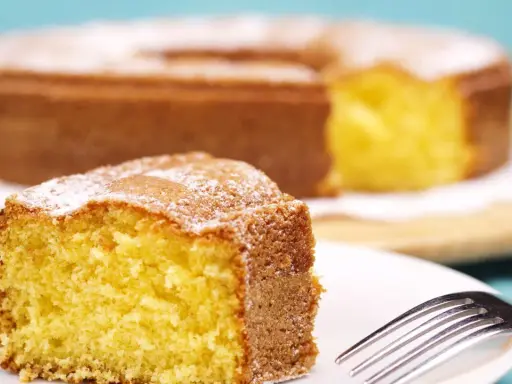 Una fetta di torta 7 vasetti soffice e dorata, cosparsa di zucchero a velo, su un piatto bianco con una forchetta.