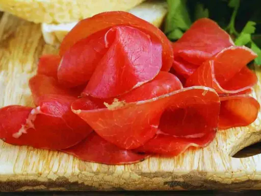 Fette sottili di bresaola, un insaccato di manzo stagionato, disposte su un tagliere di legno con pane e prezzemolo sullo sfondo.