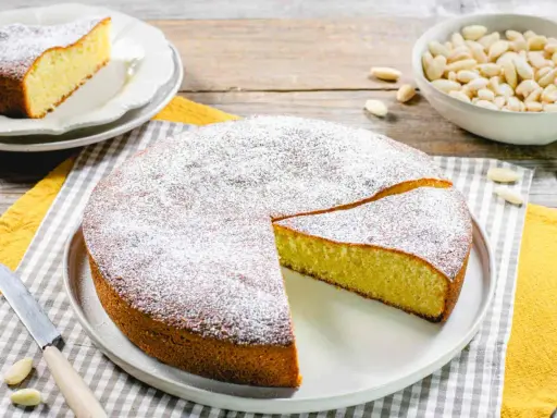 Una deliziosa torta con farina di mandorle, spolverata di zucchero a velo, pronta per essere gustata. Accanto, una ciotola di mandorle intere.