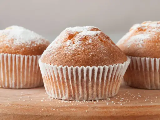 Tre deliziosi muffin senza glutine, spolverati di zucchero a velo, su un tagliere di legno.