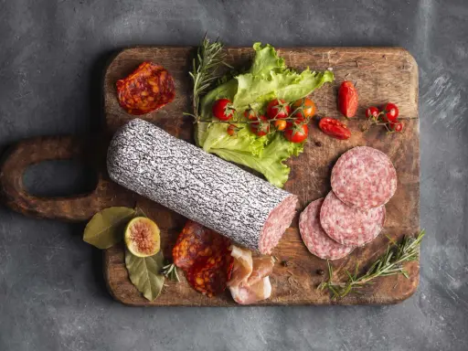 Assortimento di tipi di salame su tagliere in legno, con pomodorini, lattuga e fichi.
